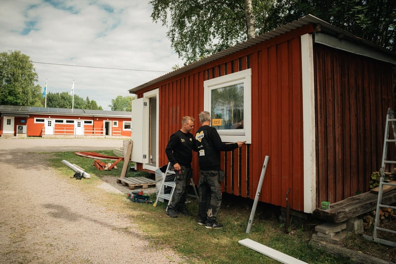 Två personer monterar in ett fönster i en röd stuga